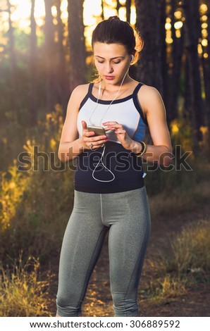 Woman listening to the music, resting after fitness in the nature at sunset. Looking at smartphone searching trough music library