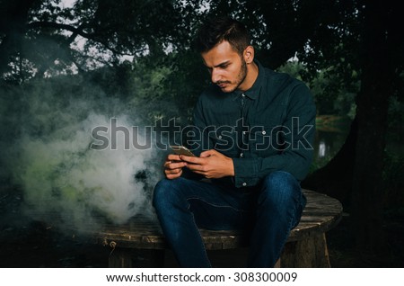 Fashionable hipster man reading a massage on his smartphone with colorful smoke bombs in nature. Vintage Instagram style effect, soft focus, low light, grain texture visible on maximum size.