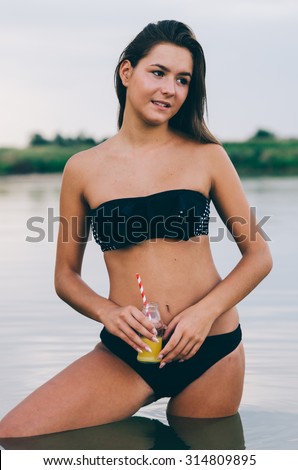 Sexy brunette in black swimsuit posing in lake water. Young female in lake during sunset holding a bottle of juice. Perfect body girl posing  in summer evening at beach.