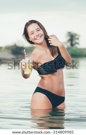 Sexy brunette in black swimsuit posing in lake water. Young female in lake during sunset holding a bottle of juice offering you to drink. Perfect body girl posing  in summer evening at beach.
