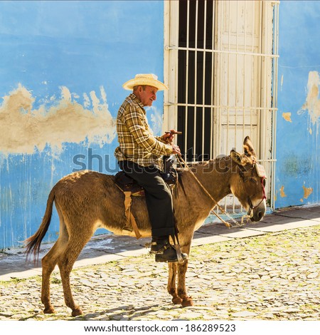 TRINIDAD, CUBA - DECEMBER 8, 2013: Old men sits on a donkey on the cobblestone street of Trinidad in Cuba and asks turists for money for a picture with him,