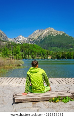 Young man in green jacket sitting on bench by clear mountains lake Strbske Pleso with High Tatra peaks in background