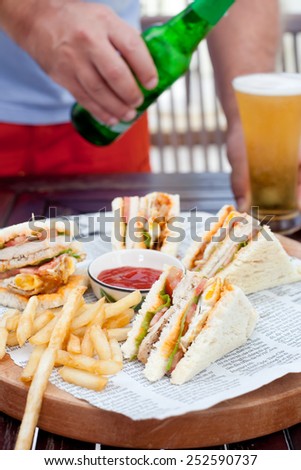 Club sandwich with french fries and a man pouring a glass of beer on a wooden table. Fast food.
