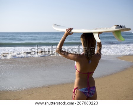 Rear view of  blonde sexy surfer young woman in bikini with white surfboard over head, wave on background