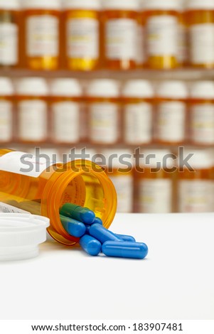 Blue capsules spilling from prescription medicine bottle on a white counter with rows of medicine bottles in a medicine cabinet in the background. Focus on opening of bottle and foreground copy space.