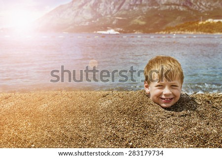 Boy buried in sand on the sea behind head smiling cloudy sky