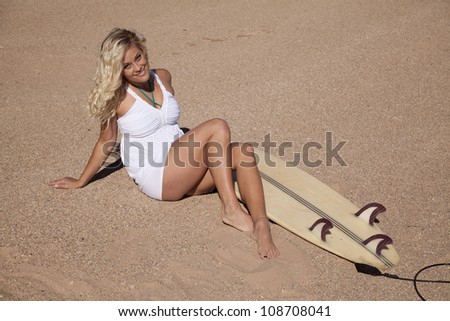 A woman is smiling and sitting by her surf board in the sand.