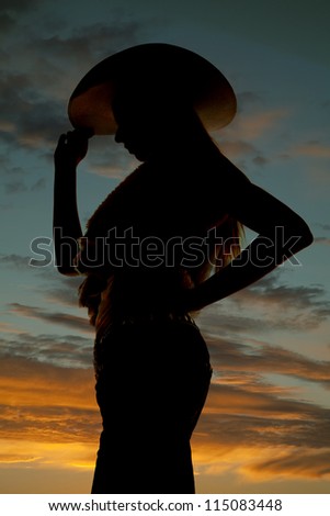 A cowgirl standing holding on to the brim of her hat with a beautiful sunset in the background.