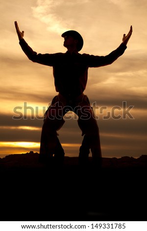 A cowboy is standing with his hands up in the air silhouetted in the sunset.