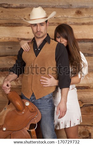 An Indian woman is hiding behind her cowboy who is holding a saddle.