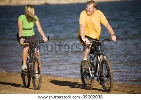 A man and a woman riding bikes together laughing and talking while riding on the beach.