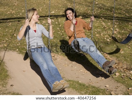 Two women laughing and smiling while swinging on the swings at the park.