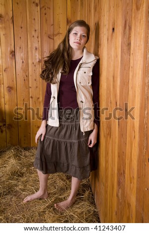 A woman standing by a wood wall with bare feet in hay, in a skirt with a serious look on her face.