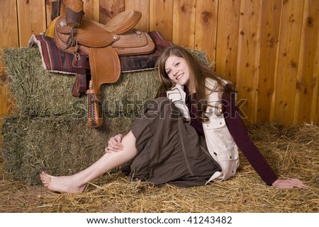 A woman sitting on the ground of hay by a saddle in a skirt with bare feet smiling.