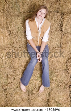 A full picture of a woman sitting on a stack of hay bales with her shoes off and a serious look on her face.