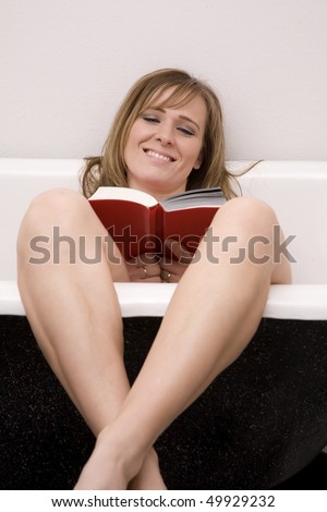 A woman sitting in her tub relaxing and enjoying her self with her legs over the side of the tub while reading a book.