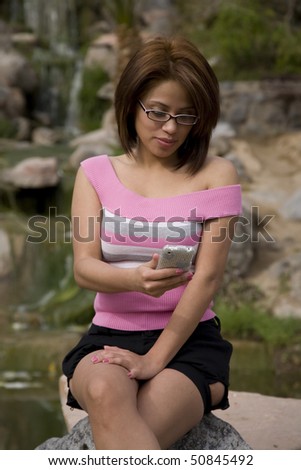 A woman sitting in the park by a pond on a rock looking at her cell phone.
