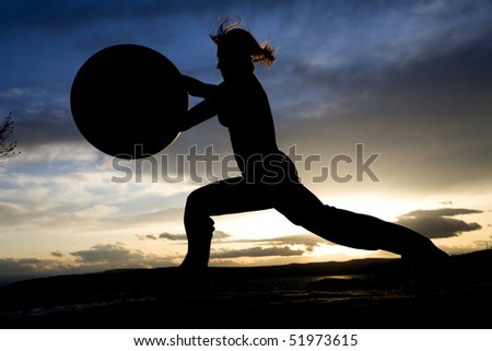 A silhouette of a woman doing a yoga pose with a exercise ball on the beach with a sunset behind her.