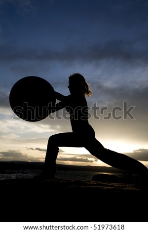 A silhouette of a woman doing a yoga pose reaching a exercise ball in the sky on the beach with a sunset behind her.
