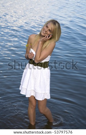 A woman in a white dress is standing in the water and talking on the cell phone.