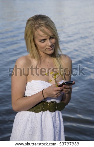 A woman in a white dress is standing in the water and texting on the cell phone.