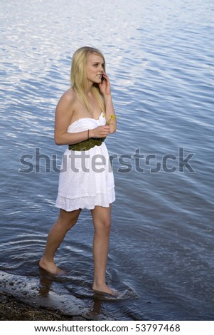A woman in a white dress walking in the water and talking on her cell phone.
