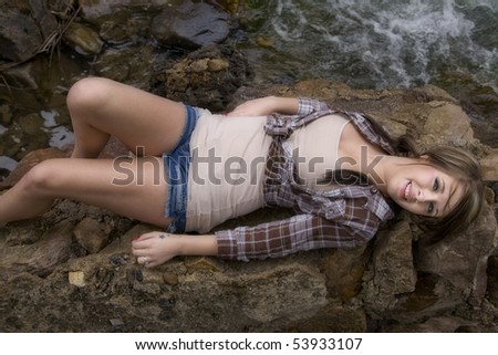A woman is laying on her back on a big rock with the river right by her.
