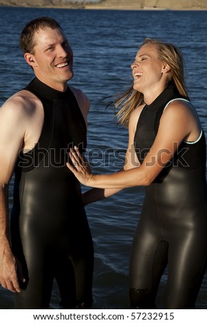 A couple that is in love laughing and smiling while standing in their wet suits in the water.
