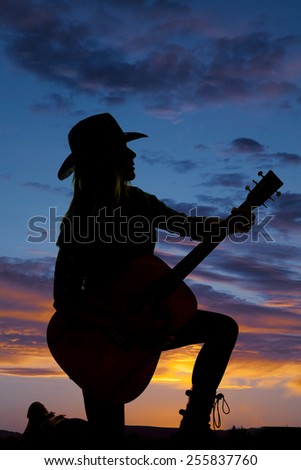 A silhouette of a woman kneeling on the ground in the outdoors holding on to her guitar.