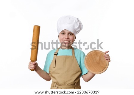 Adorable Caucasian teenage boy - baker wearing chef uniform, smiles a cheerful toothy smile, posing to camera with a wooden cutting board and rolling pin in his hands, isolated over white background Foto stock © 