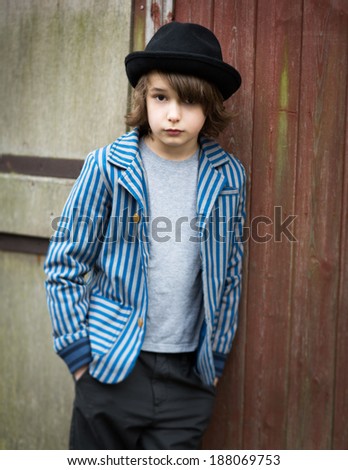 Portrait of a boy in a striped jacket and hat leaning against a wood cladded wall.