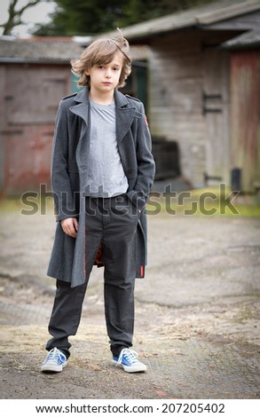 Portrait of a boy with long hair in a long coat standing isolated in a farm yard in front of stables hair blowing in the wind