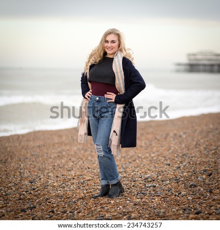 Portrait of a beautiful blond teenage girl with long hair wearing denim jeans, woolly jumper, long coat and a scarf standing alone on a cold pebble beach with her hands placed on her hips.