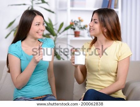 Two happy young female friends with coffee cups conversing in the living room at home