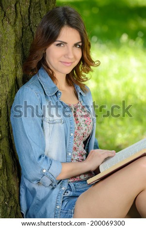 Beautiful young woman-student reading book on grass, against background of summer green park.