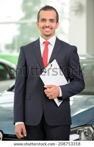 Handsome young car salesman standing at the dealership and pointing car