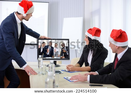 Group of male and female businesspeople seated at a table watching an online conference on a computer screen, during the celebration of Christmas