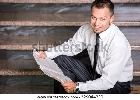 Young unemployed man is looking for a job in the newspaper, sitting on the stairs.