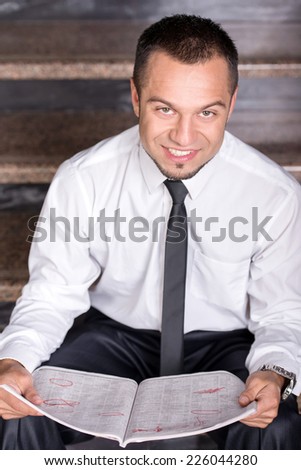 Young unemployed man is looking for a job in the newspaper, sitting on the stairs. Looking at the camera.