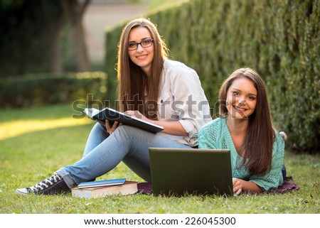 Two smiling female students are sitting on the grass with laptop at campus. They are looking at camera.