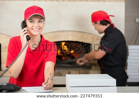 Smiling delivery woman of pizza on the phone and chef baker with long handled bread pan baking pizza into wood fire oven done.