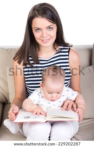 Beautiful woman is holding her baby and a book in her arms while sitting on a sofa.