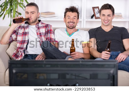 Three young football fans are cheering football match at home and drinking beer.