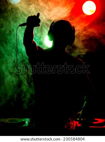 Silhouette of a young DJ at work with a microphone and the club lights on the background.