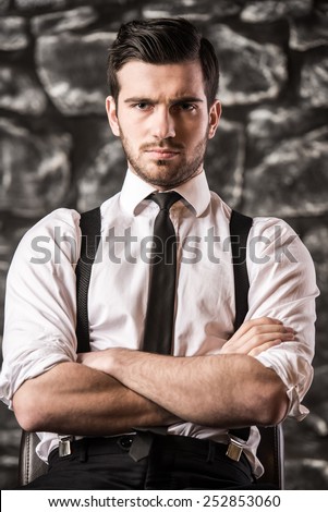 Confident young man in shirt and suspenders is sitting on the chair and looking at you.