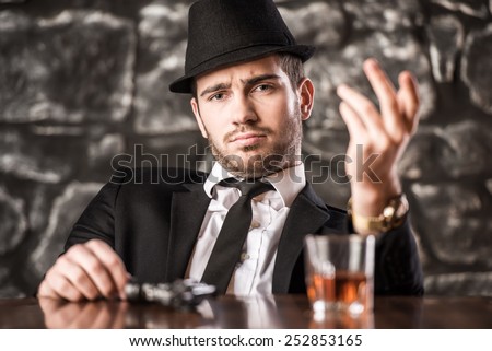 Confident, gangster man in suit and hat is sitting at the table with a glass of whisky and gun.