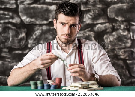 Confident poker player. Serious young man in shirt and suspenders is  sitting at the poker table and holding money.