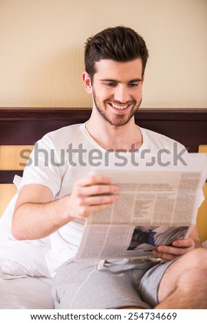 Young, smiling man is reading a newspaper, sitting on bed at morning.
