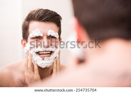 Shaving with fun. Rear view of playful young man with shaving cream on his face is standing in front of the mirror and smiling.