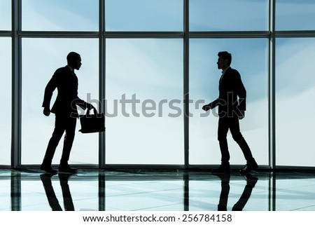 Silhouette view of two young businessmen are standing in modern office with panoramic windows.